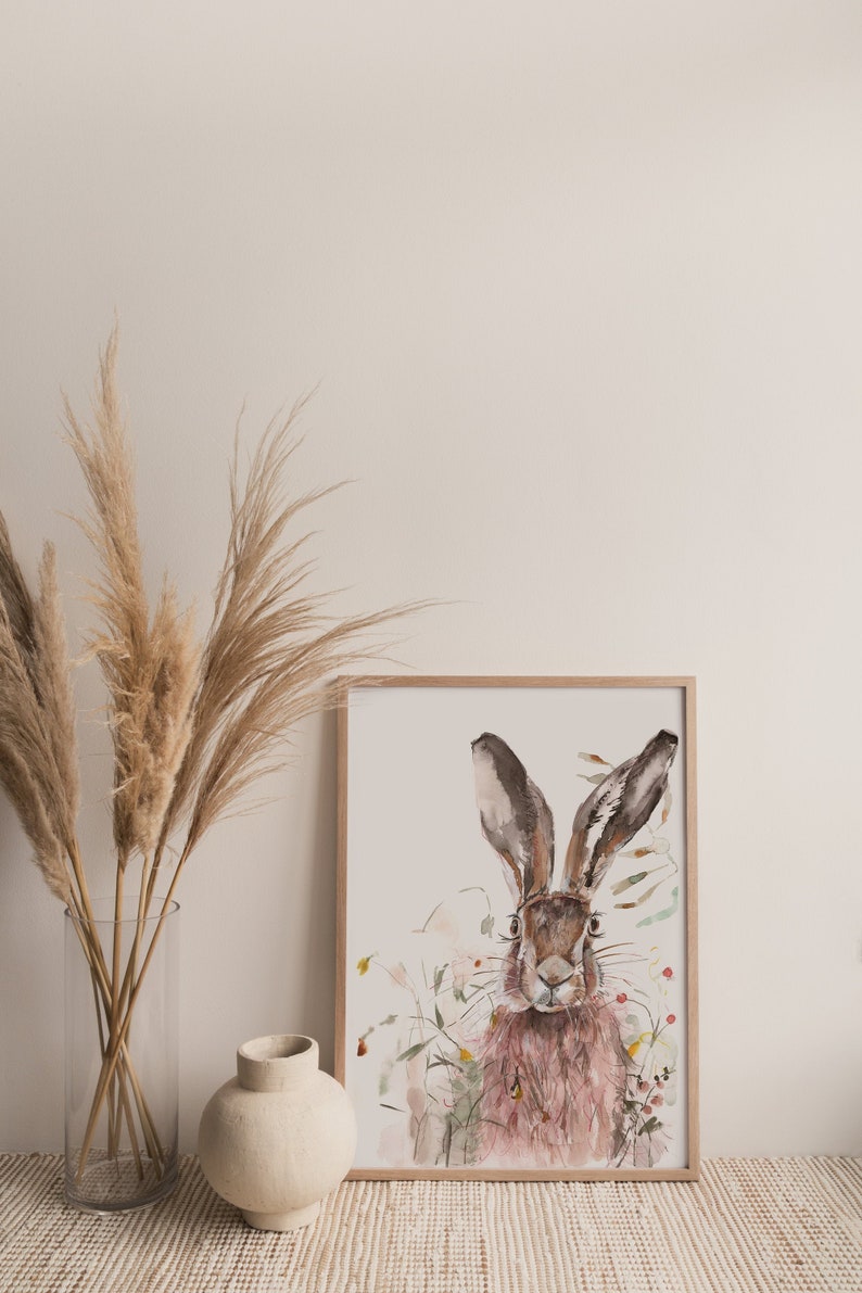 May include: A watercolour painting of a brown hare with long ears, surrounded by pink and green wildflowers. The painting is framed in a light wood frame and is displayed on a white wall with a vase of dried pampas grass.