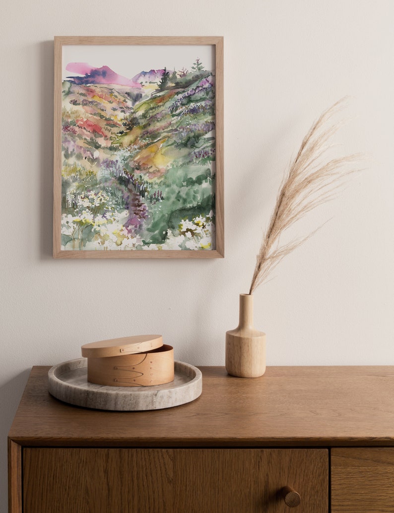 May include: A framed watercolor painting of a green, yellow, and purple landscape with a winding path. The painting is hanging on a white wall above a wooden dresser with a small wooden box and a vase with dried grass.