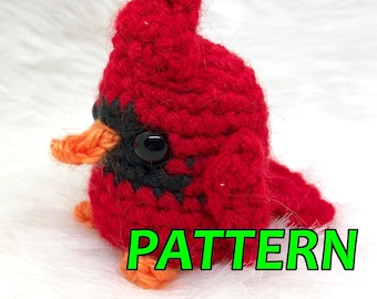 Puede incluir: Un pájaro cardenal rojo tejido a ganchillo, con detalles negros y un pico naranja. El pájaro tiene un ojo negro grande y está etiquetado "PATTERN" en texto verde. Probablemente sea un patrón de manualidades.
