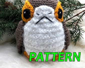Puede incluir: Un juguete de peluche tejido a crochet que se asemeja a un personaje de Star Wars. El juguete es marrón, blanco y naranja con ojos negros. El juguete está sentado sobre una superficie blanca y esponjosa con ramas de pino verdes en el fondo. La palabra "PATTERN" está escrita en letras verdes en la parte inferior de la imagen.