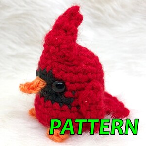 May include: A handmade, crocheted red cardinal bird with black accents and an orange beak. The bird has a large, black eye and is labeled "PATTERN" in green text. This item is likely a craft pattern.