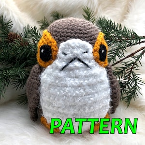 May include: A crocheted stuffed toy resembling a character from Star Wars. The toy is brown, white, and orange with black eyes. The toy is sitting on a white fluffy surface with green pine branches in the background. The word "PATTERN" is written in green letters at the bottom of the image.