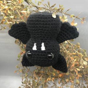 Puede incluir: Un murci&eacute;lago de crochet negro con ojos y dientes bordados en blanco. El murci&eacute;lago est&aacute; colgado de una rama.