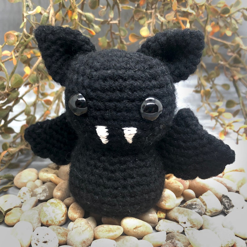 Puede incluir: Un murci&eacute;lago de crochet negro con dientes blancos y grandes ojos negros. El murci&eacute;lago est&aacute; sentado en una cama de peque&ntilde;as piedras de color marr&oacute;n claro.