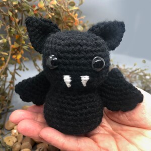 Puede incluir: Un murci&eacute;lago de crochet negro con dientes blancos y ojos negros de bot&oacute;n. El murci&eacute;lago est&aacute; sentado en la mano de una persona.