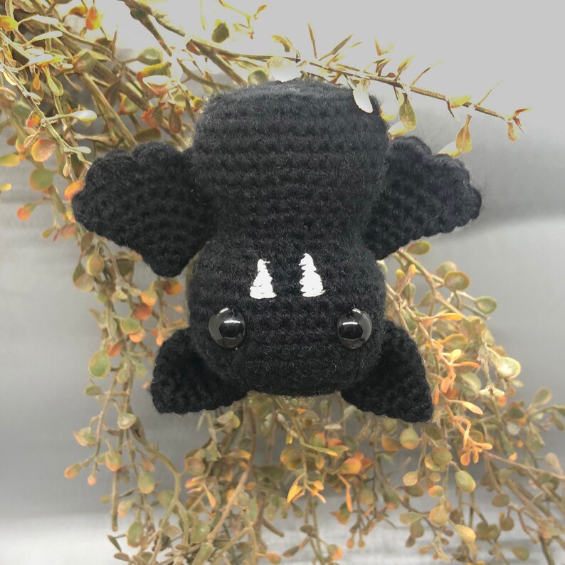 Puede incluir: Un murci&eacute;lago de crochet negro con ojos blancos y dientes blancos colgando de una rama de hojas verdes.