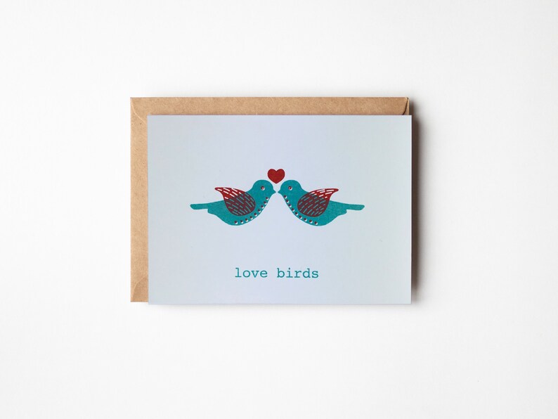 May include: A greeting card with a light blue background featuring two teal blue birds facing each other with a red heart above them. The text "love birds" is printed below the birds.