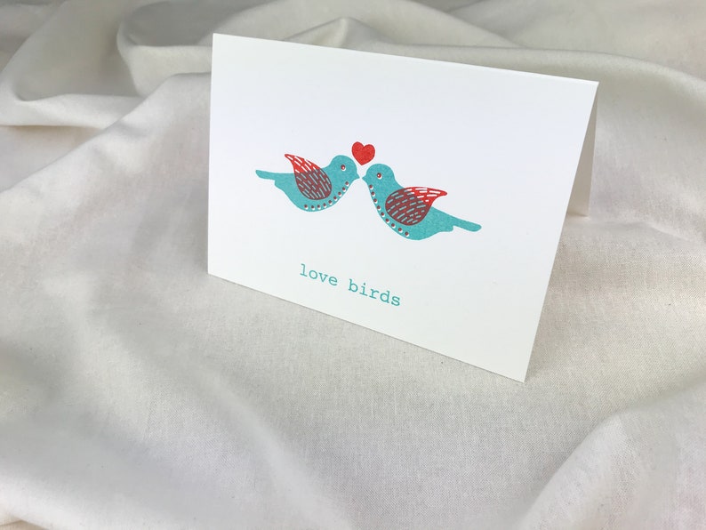 May include: A greeting card with two blue birds kissing with a red heart above them. The text "love birds" is printed below the birds.