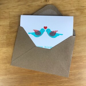 May include: A brown envelope with a white card inside. The card features two blue birds with red accents, facing each other and kissing. The text "love birds" is visible at the bottom of the card.
