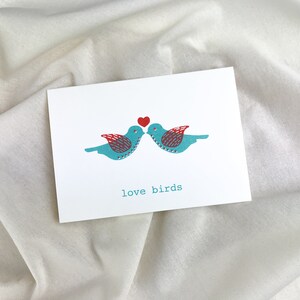 May include: A white card with two blue birds facing each other with a red heart above them. The text "love birds" is printed below the birds.