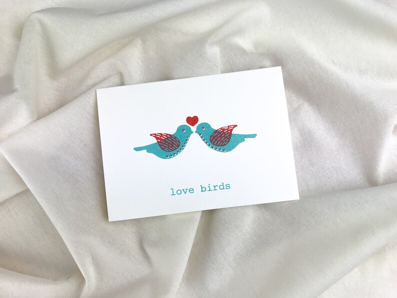 May include: A white card with two blue birds facing each other with a red heart above them. The text "love birds" is printed below the birds.