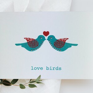 May include: A white card with two teal blue birds facing each other with a red heart above them. The text "love birds" is printed below the birds.