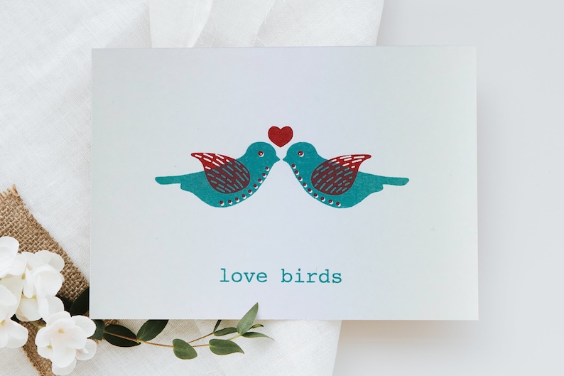May include: A white card with two teal blue birds facing each other with a red heart above them. The text "love birds" is printed below the birds.