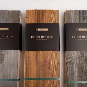 Pode incluir: Três pacotes de tábuas de madeira rústica em tons de cinza e castanho. Cada pacote é embrulhado com uma faixa preta com o texto "DECOR RUSTICO THE ART OF WOOD" em letras douradas. Uma fita verde prende cada pacote.