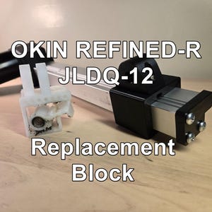 May include: A close-up of a replacement block for an OKIN REFINED-R JLDQ-12 mechanism. The block is black and silver, with the text "Replacement Block" in white. The image is on a light brown surface.