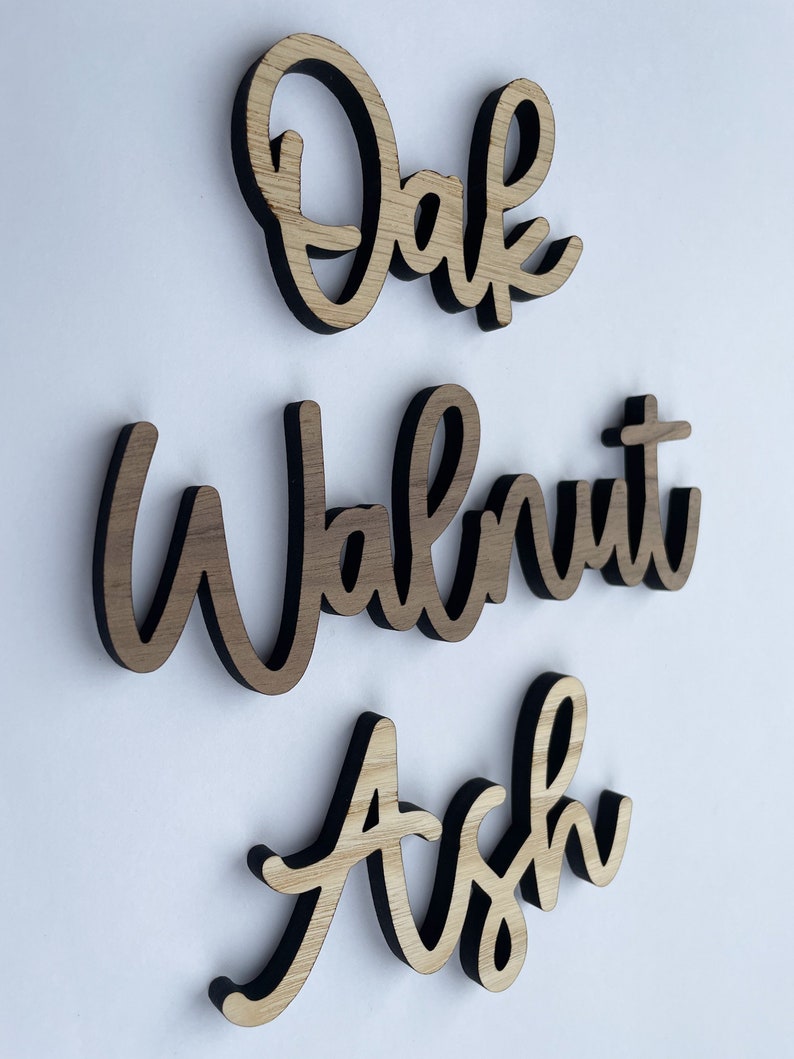 May include: Three wooden signs with the words "Oak", "Walnut", and "Ash" written in a cursive font. The signs are made of light brown wood with a dark brown border.