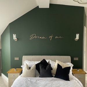 May include: A bedroom with a dark green wall and a white bed with black and white pillows. The wall has a wooden sign that says "Dream of me."