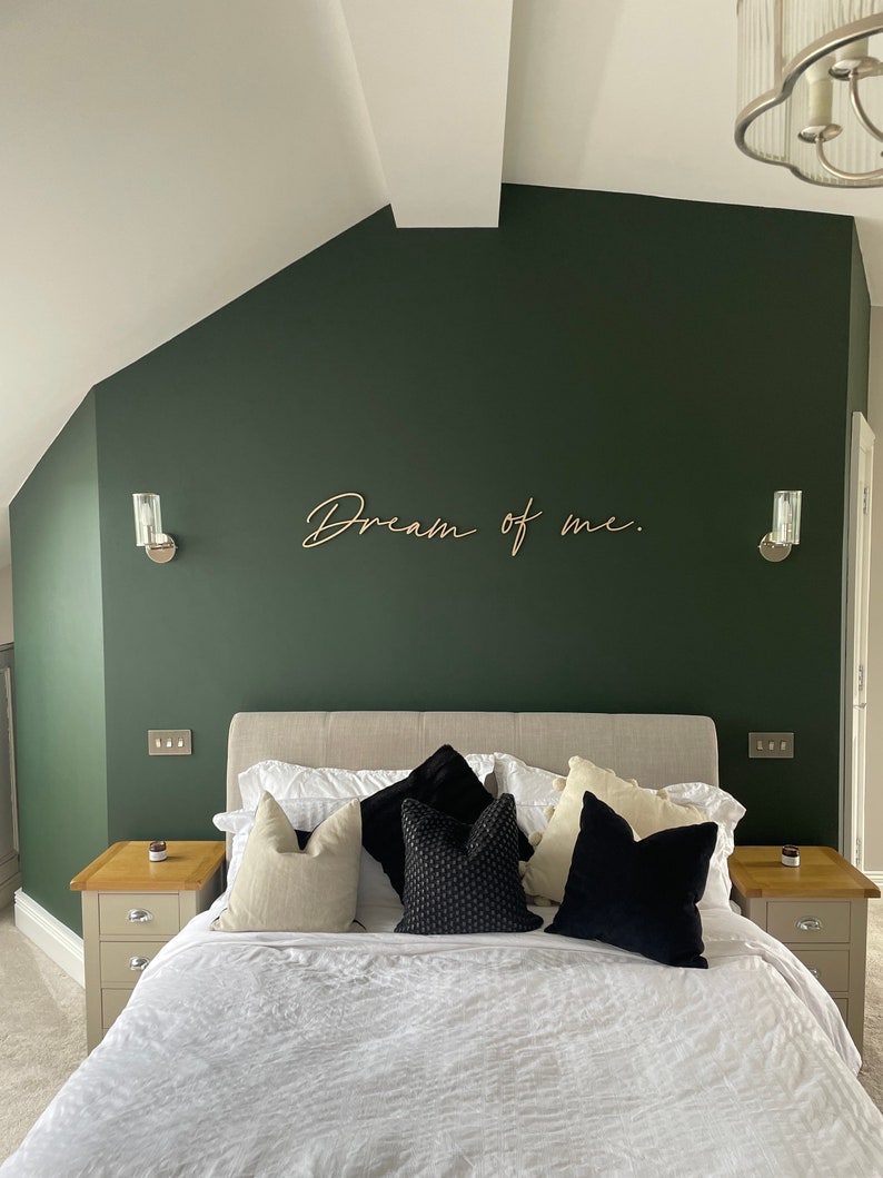 May include: A bedroom with a dark green wall and a white bed with black and white pillows. The wall has a wooden sign that says "Dream of me."