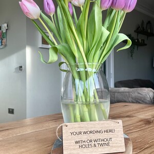 May include: Wooden sign with the text "YOUR WORDING HERE! WITH OR WITHOUT HOLES & TWINE" in black lettering. The sign is hanging from a piece of twine and is sitting on a wooden surface. A vase of pink and purple tulips is in the background.