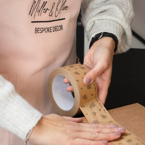 May include: A person is using a brown paper tape dispenser with a white core and a printed design of leaves and the text "100% RECYCLABLE" to seal a brown cardboard box.