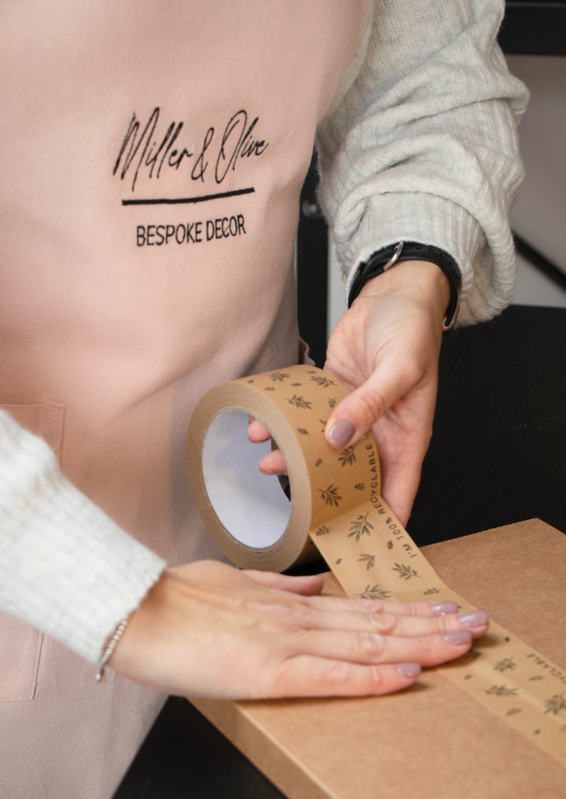 May include: A person is using a brown paper tape dispenser with a white core and a printed design of leaves and the text "100% RECYCLABLE" to seal a brown cardboard box.