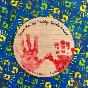 May include: A wooden circle with red handprints and the text "You're the best Daddy, hands down! Love from Ezra & Otis x Father's Day 2022".