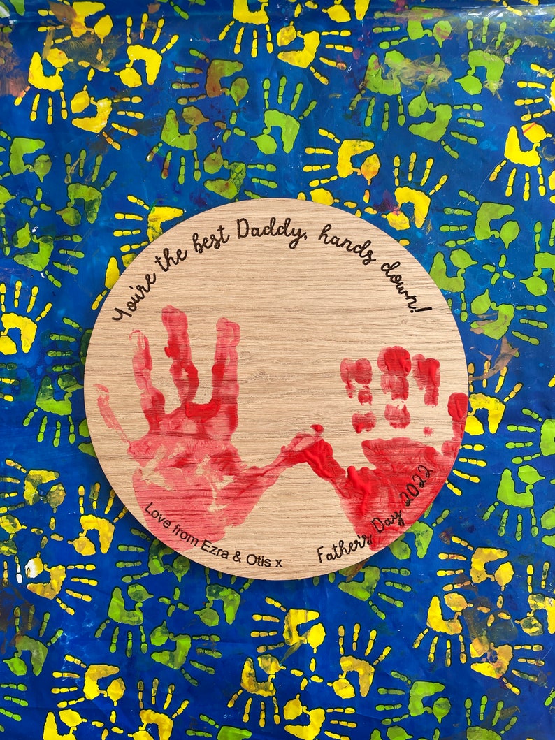 May include: A wooden circle with red handprints and the text "You're the best Daddy, hands down! Love from Ezra & Otis x Father's Day 2022".