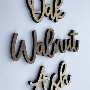 May include: Three wooden signs with the words "Oak", "Walnut", and "Ash" written in a cursive font. The signs are made of light brown wood with a dark brown border.