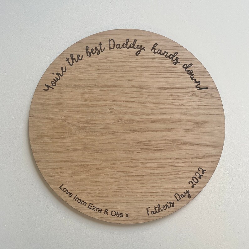 May include: A round wooden plaque with the text "You're the best Daddy, hands down!" and "Love from Ezra & Otis x Father's Day 2022" engraved on it.