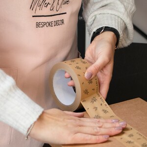May include: A person is using a brown tape dispenser with a white core and a printed design of leaves and the text "100% RECYCLABLE" to seal a brown cardboard box. The tape dispenser has a black handle.