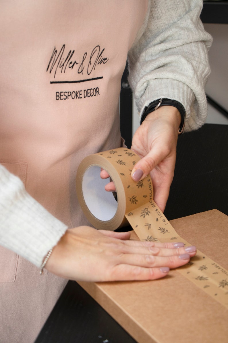 May include: A person is using a brown tape dispenser with a white core and a printed design of leaves and the text "100% RECYCLABLE" to seal a brown cardboard box. The tape dispenser has a black handle.