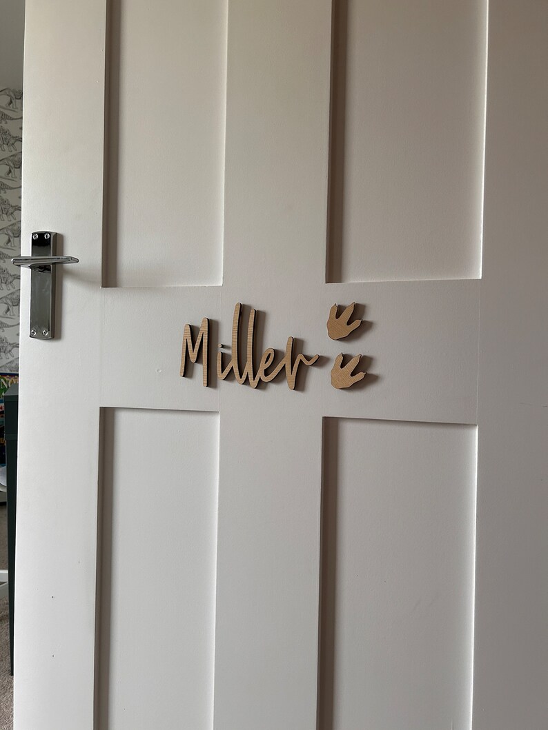 May include: A white door with a wooden name sign that says "Miller" with two dinosaur footprints on the right side of the name.
