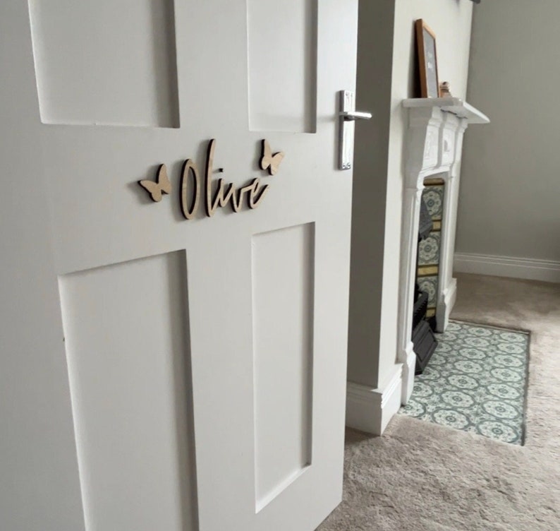 May include: A white door with a wooden name sign that says "Olive" with two butterfly cutouts on either side of the name.