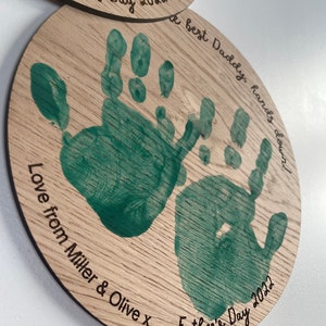 May include: A wooden round plaque with two green handprints and the text "A best Daddy, hands down" and "Love from Miller & Olive x Father's Day 2022".