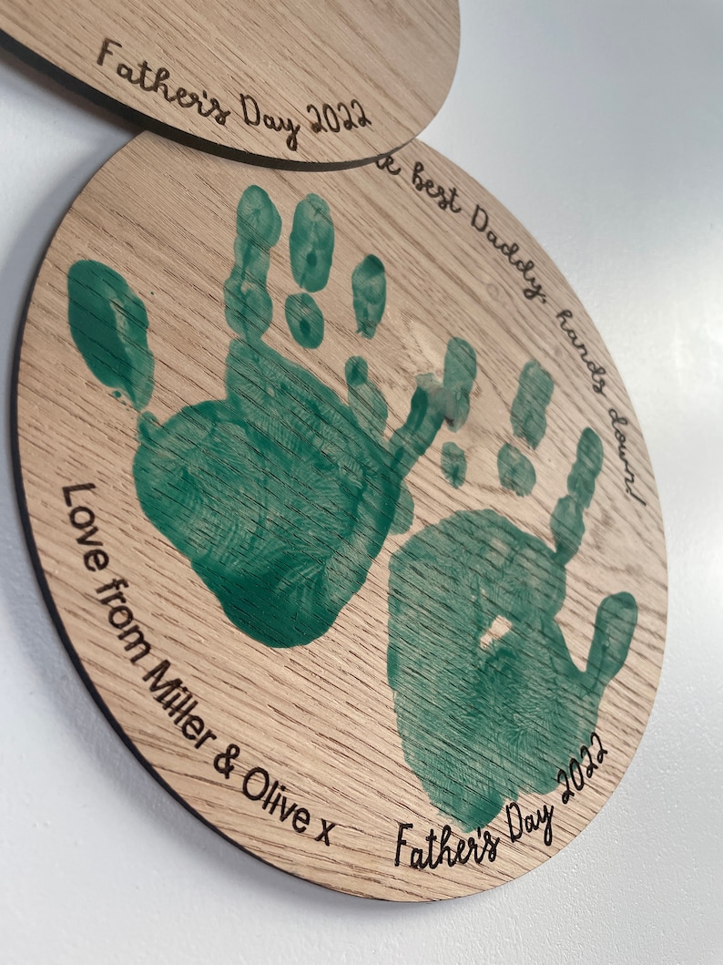May include: A wooden round plaque with two green handprints and the text "A best Daddy, hands down" and "Love from Miller & Olive x Father's Day 2022".