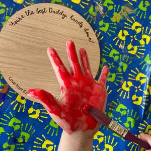 May include: A wooden round plaque with the text "You're the best Daddy, hands down! Love from Ez" engraved on it. A child's hand is covered in red paint and a paintbrush is being used to apply more paint.