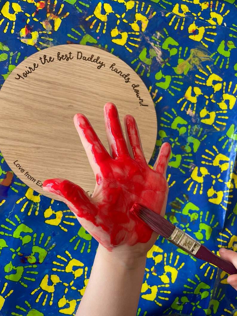 May include: A wooden round plaque with the text "You're the best Daddy, hands down! Love from Ez" engraved on it. A child's hand is covered in red paint and a paintbrush is being used to apply more paint.