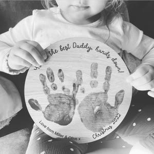 May include: A round wooden plaque with two handprints in black and white. The plaque has the text "You are the best Daddy, hands down! Love from Miller & Olive x Christmas 2022" engraved on it.