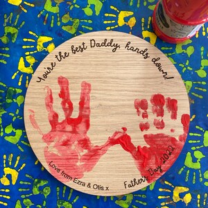 May include: A wooden round plaque with two red handprints and the text "You're the best Daddy, hands down! Love from Ezra & Otis x Father's Day 2022"