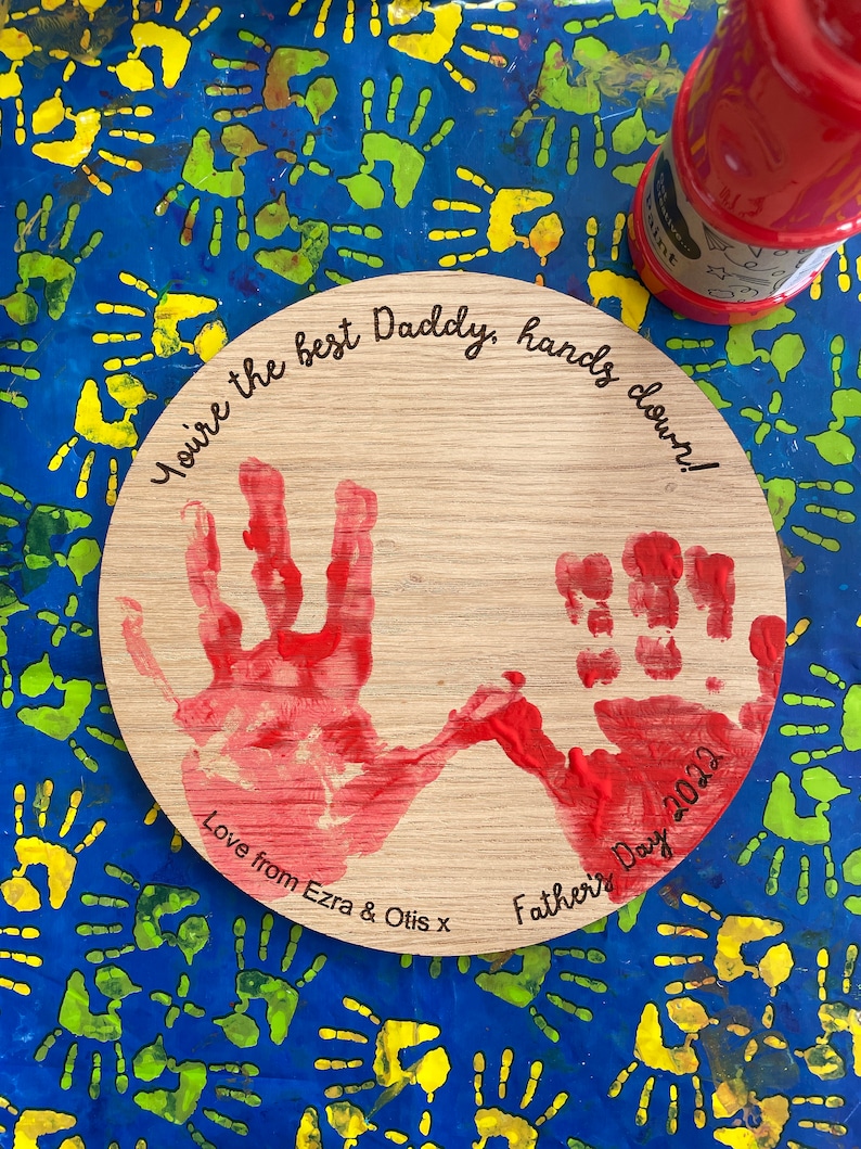 May include: A wooden round plaque with two red handprints and the text "You're the best Daddy, hands down! Love from Ezra & Otis x Father's Day 2022"