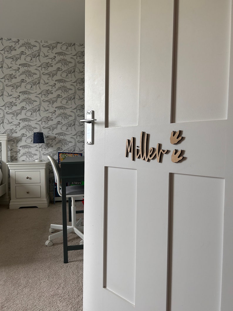 May include: A white door with a wooden nameplate that reads "Miller" with two small wooden dinosaur footprints on the right side of the name.