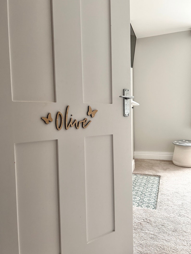 May include: A white door with a wooden name sign that says "Olive" with two butterfly cutouts on either side of the name.