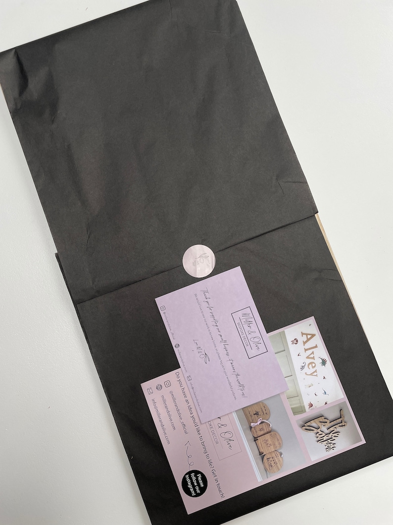 May include: A pink and white business card with the text "Miller & Olive Bespoke Decor" and the text "Thank you for supporting my small business!"  The card also includes a website address, Instagram handle, and email address. The card is sitting on top of a black piece of tissue paper.