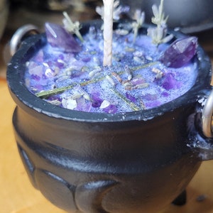 May include: A black cauldron-shaped candle with a purple wax interior. The candle is decorated with amethyst crystals, lavender sprigs, and glitter. A white wick rises from the center. The cauldron has three legs and two metal handles.