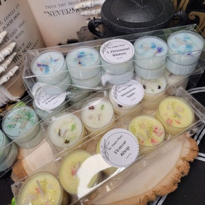 May include: A clear plastic box filled with teal and yellow scented tea light candles. The candles have dried flowers and herbs embedded in the wax. The labels on the candles read "My Magic Place", "A Thousand Wishes", "Cinnamon Apple", and "Flower Shop".