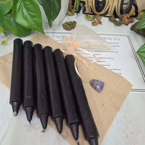 May include: Six solid black taper candles arranged in a row on a brown paper sheet. A small, sheer peach-colored drawstring pouch sits next to the candles, containing a single polished, triangular purple amethyst gemstone with visible white veining.