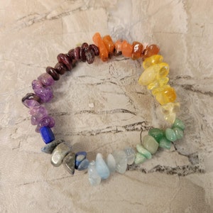 May include: A colorful bracelet made of polished, irregularly shaped gemstones. The stones are arranged in a rainbow spectrum, transitioning from purple to red, orange, yellow, green, blue, and light blue. The bracelet is on a neutral, textured surface.