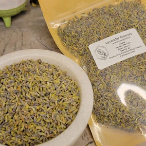 May include: A close-up of a bag of dried lavender flowers. The bag is labeled "Lavender Flowers" and lists the plant's magical properties, including its gender, planet, element, and powers. The flowers are a light purple color and have a delicate, fragrant aroma.