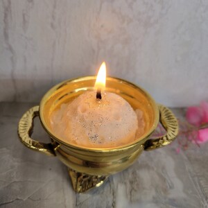 May include: A lit, round candle with glitter sits in a gold-colored bowl with handles. The candle flame is bright against the soft, textured candle. The bowl has a decorative base and is set against a neutral background.