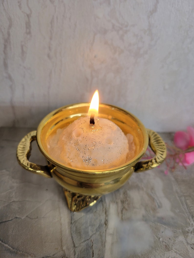 May include: A lit, round candle with glitter sits in a gold-colored bowl with handles. The candle flame is bright against the soft, textured candle. The bowl has a decorative base and is set against a neutral background.